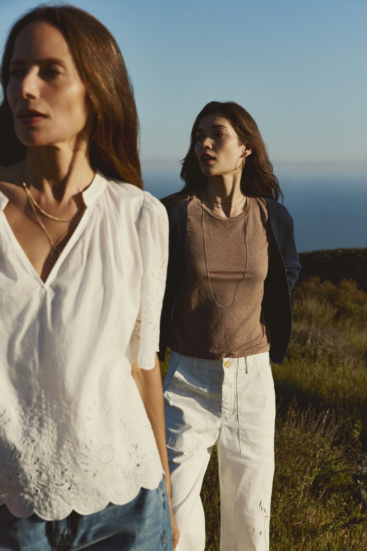 Two women standing in a field overlooking the sea, one in a white blouse and Velvet by Graham & Spencer MYA Cotton Canvas Pant, the other in a brown top and white pants.-37535283380417