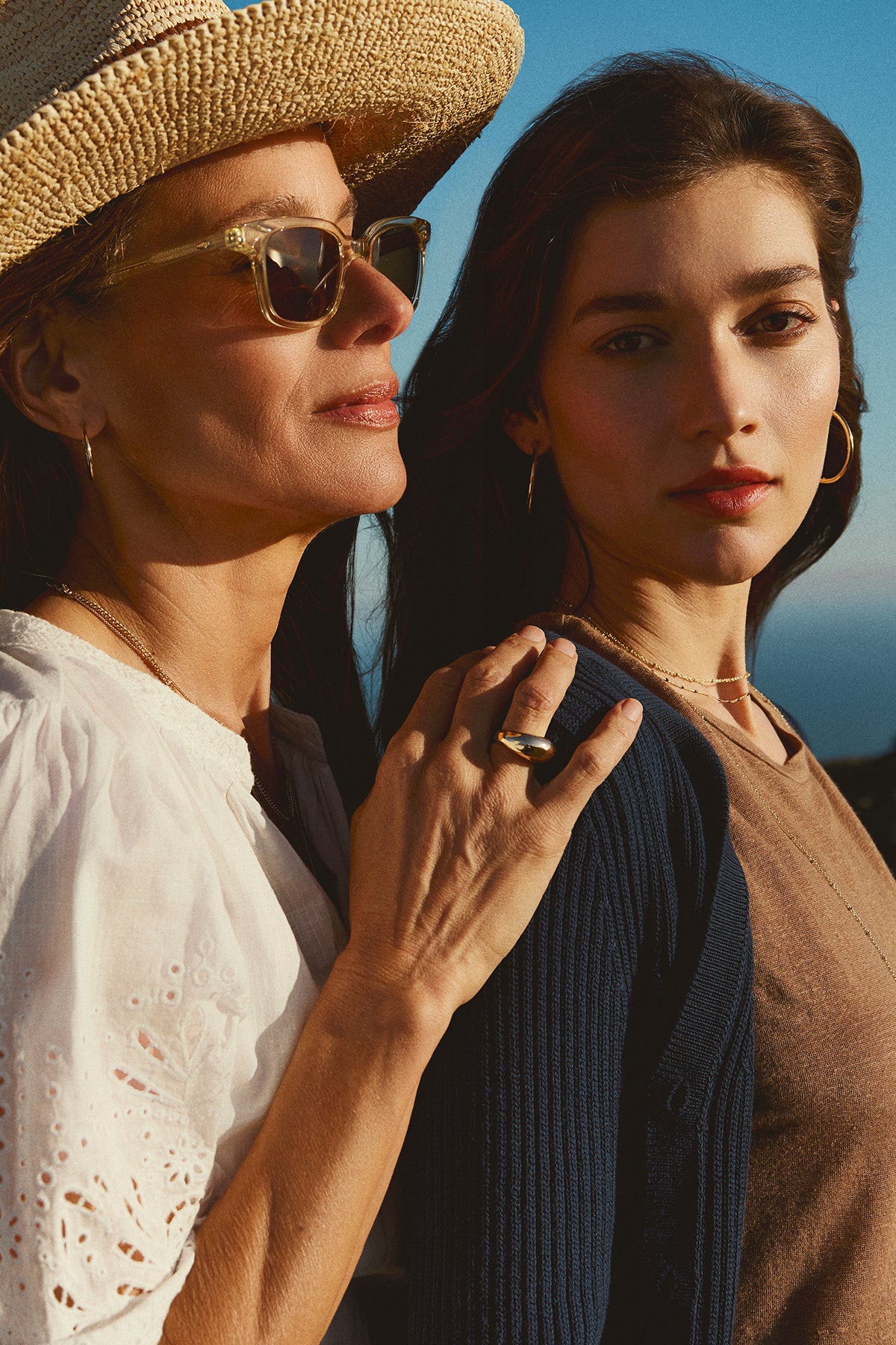   Two women standing close together, one wearing a straw hat and sunglasses, the other in a HYDIE BUTTON FRONT CARDIGAN from Velvet by Graham & Spencer, both looking towards the camera. 