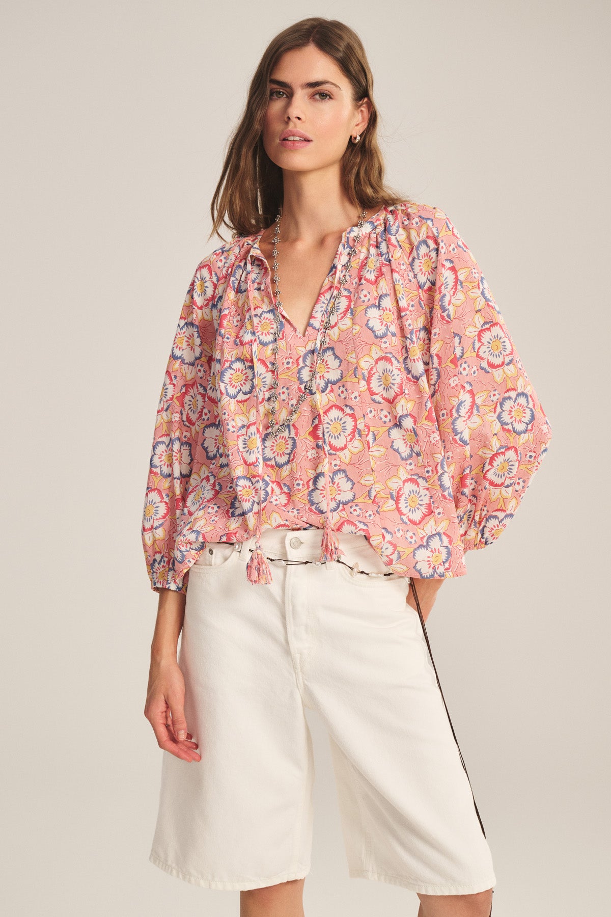 A woman wears the REESE TOP by Velvet by Graham & Spencer—a loose, lightweight cotton V-neck with a pink floral print and tassels—paired with white knee-length shorts, standing against a plain background.-41417932996801