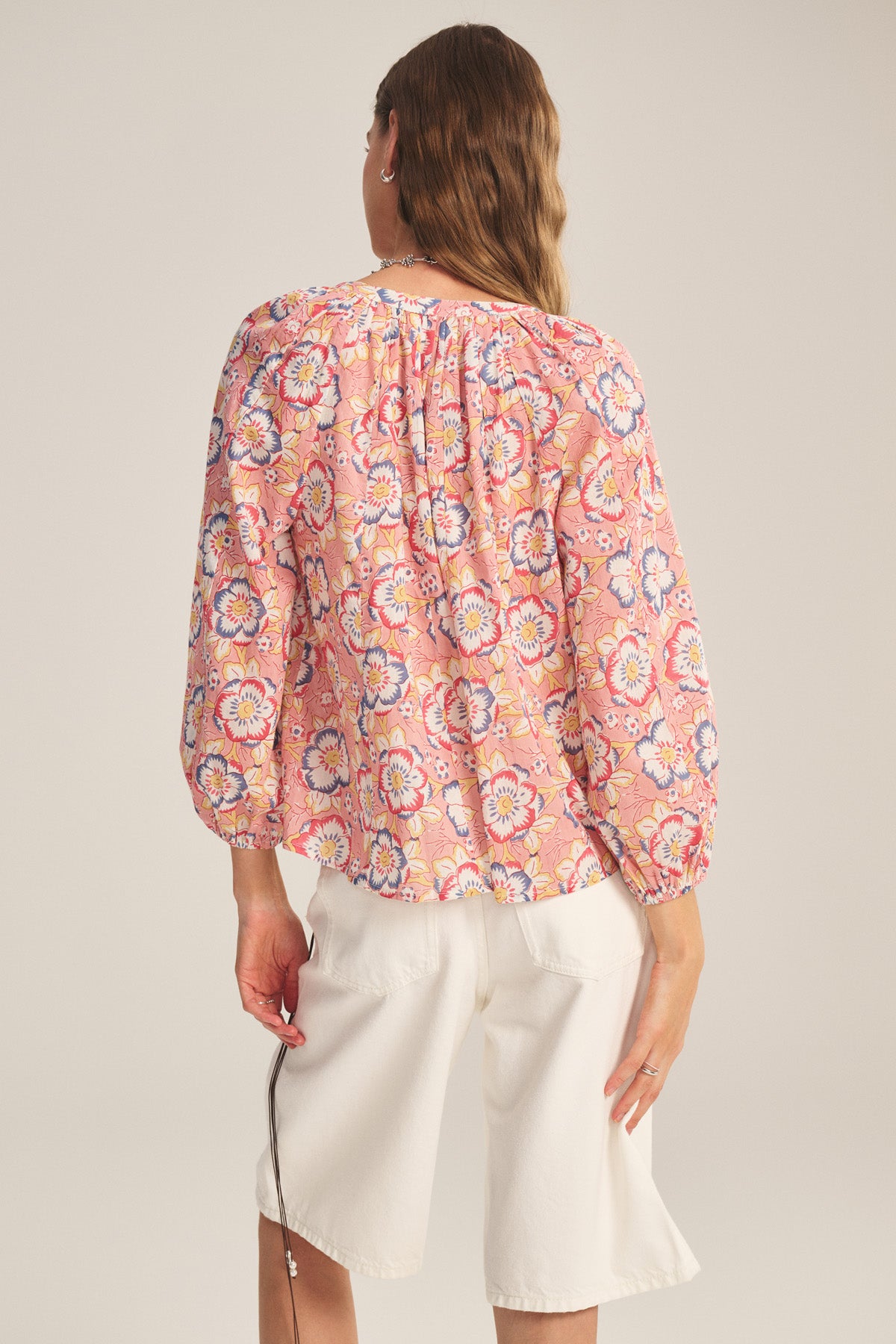 A person stands facing away, wearing the REESE TOP by Velvet by Graham & Spencer—a pink floral lightweight cotton top with puffed sleeves—paired with white knee-length shorts, against a plain light background.
