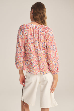 A person stands facing away, wearing the REESE TOP by Velvet by Graham & Spencer—a pink floral lightweight cotton top with puffed sleeves—paired with white knee-length shorts, against a plain light background.