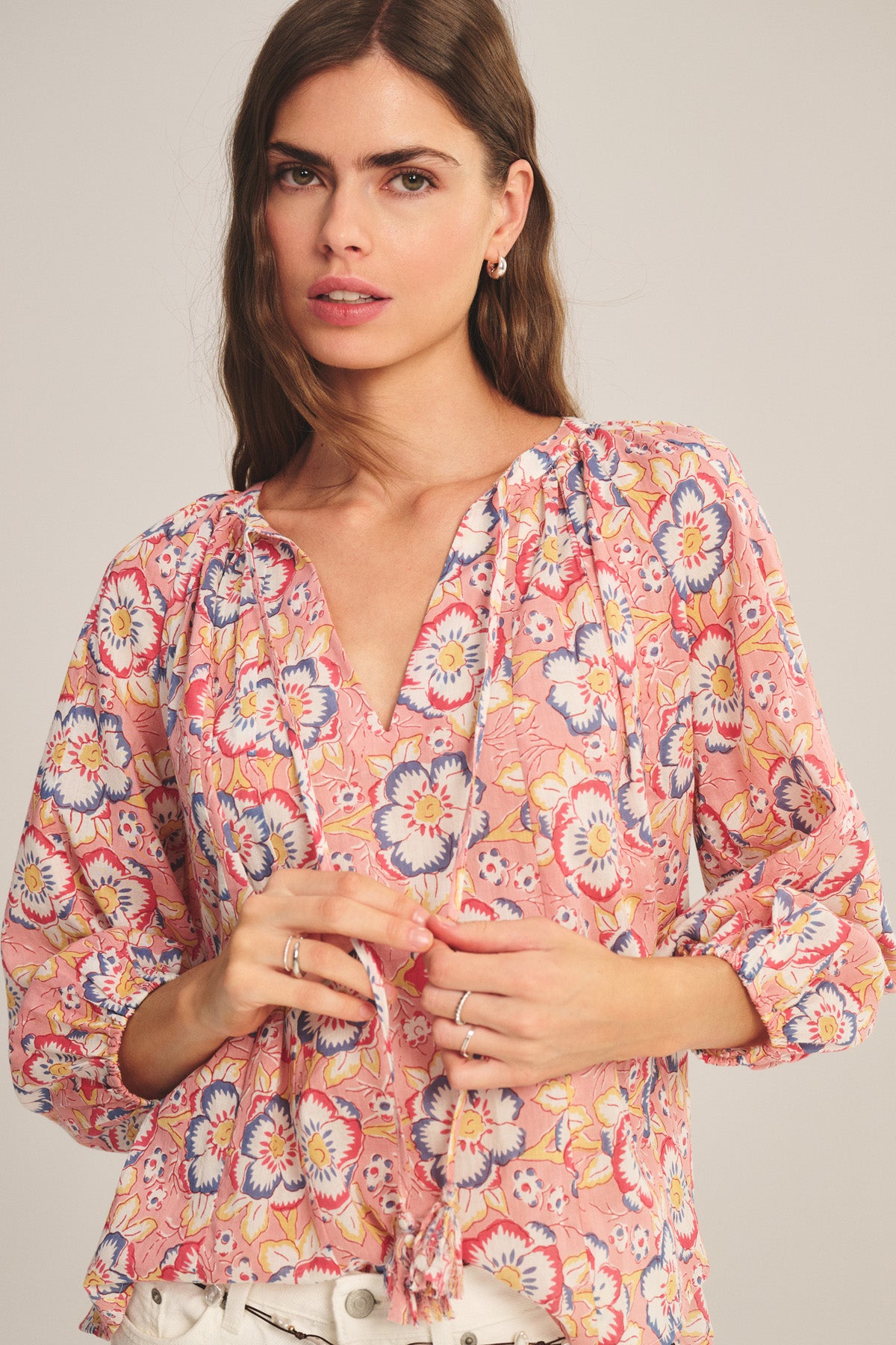 A woman stands facing the camera in the Velvet by Graham & Spencer REESE TOP—a lightweight cotton blouse with pink floral print, puffed sleeves, and V-neckline—paired with white pants, holding its drawstrings in her hands.