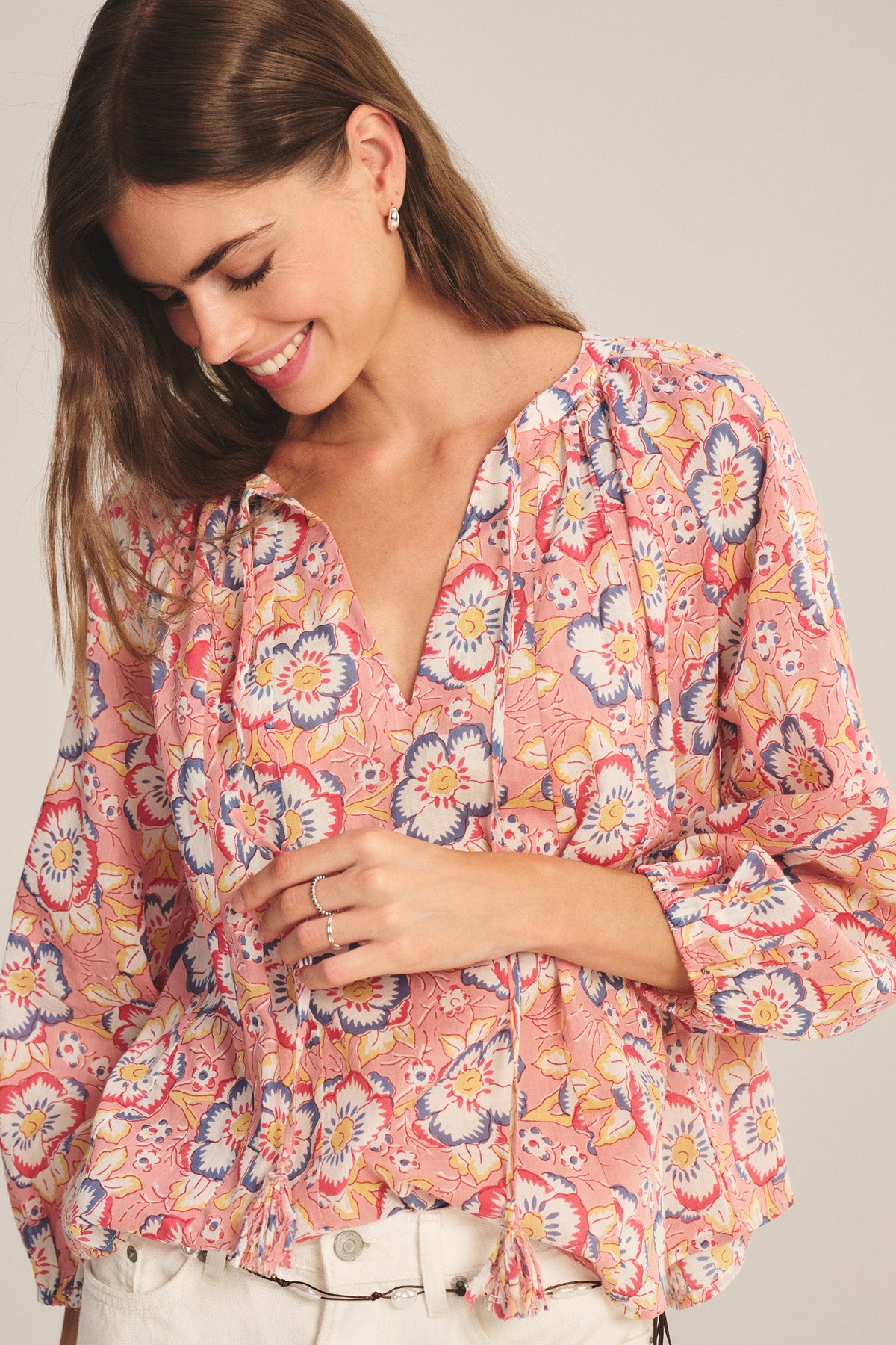 A woman smiles and looks down wearing the REESE TOP by Velvet by Graham & Spencer—a pink floral lightweight cotton V-neck top with tassels—paired with white pants and minimal jewelry.-41417933127873