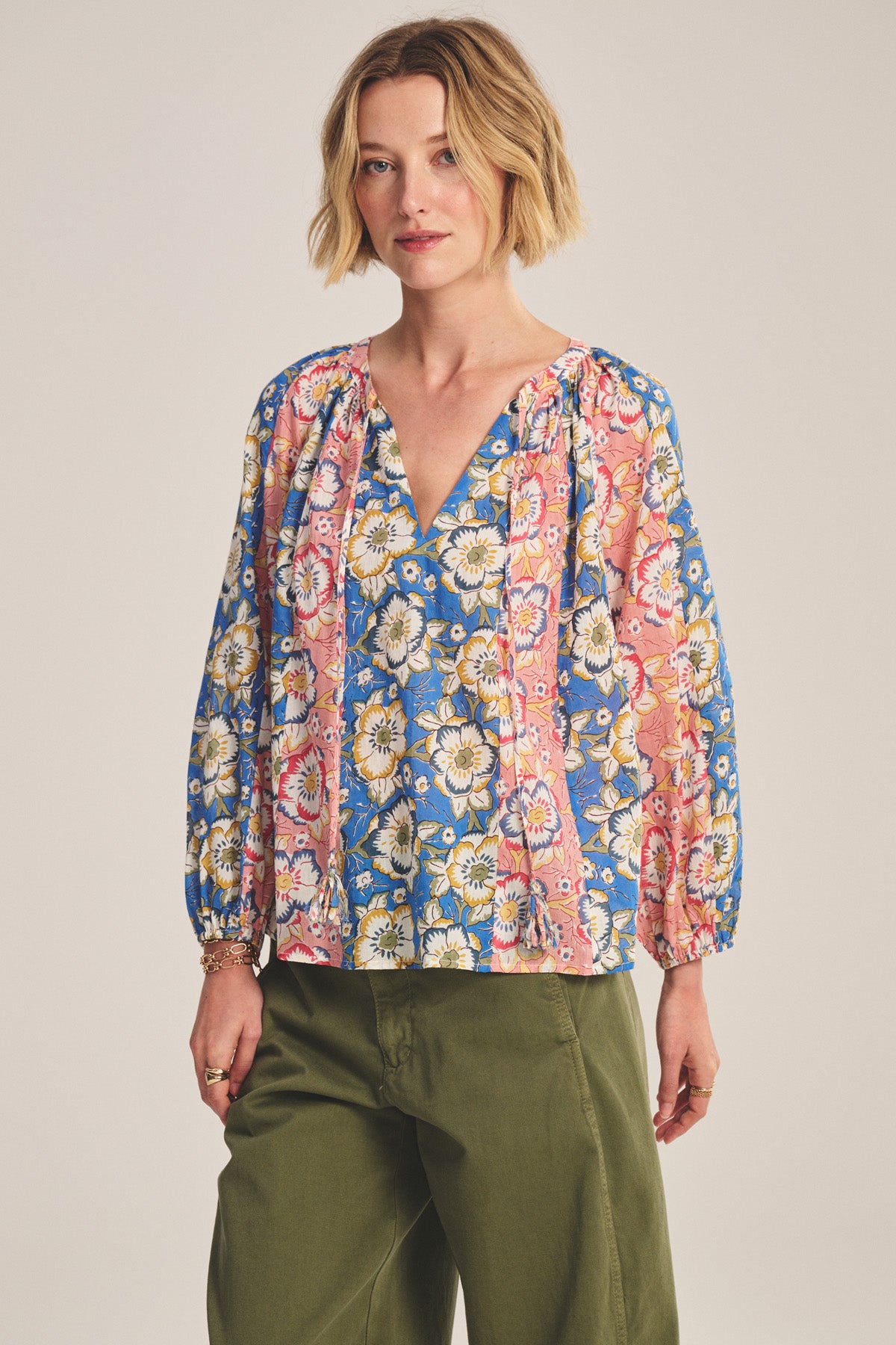 A woman with short blonde hair wears the REESE TOP by Velvet by Graham & Spencer, featuring a lightweight cotton fabric with a colorful floral pattern, paired with olive green pants, standing against a neutral background.