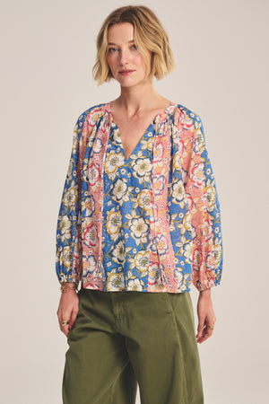 A woman with short blonde hair wears the REESE TOP by Velvet by Graham & Spencer, featuring a lightweight cotton fabric with a colorful floral pattern, paired with olive green pants, standing against a neutral background.