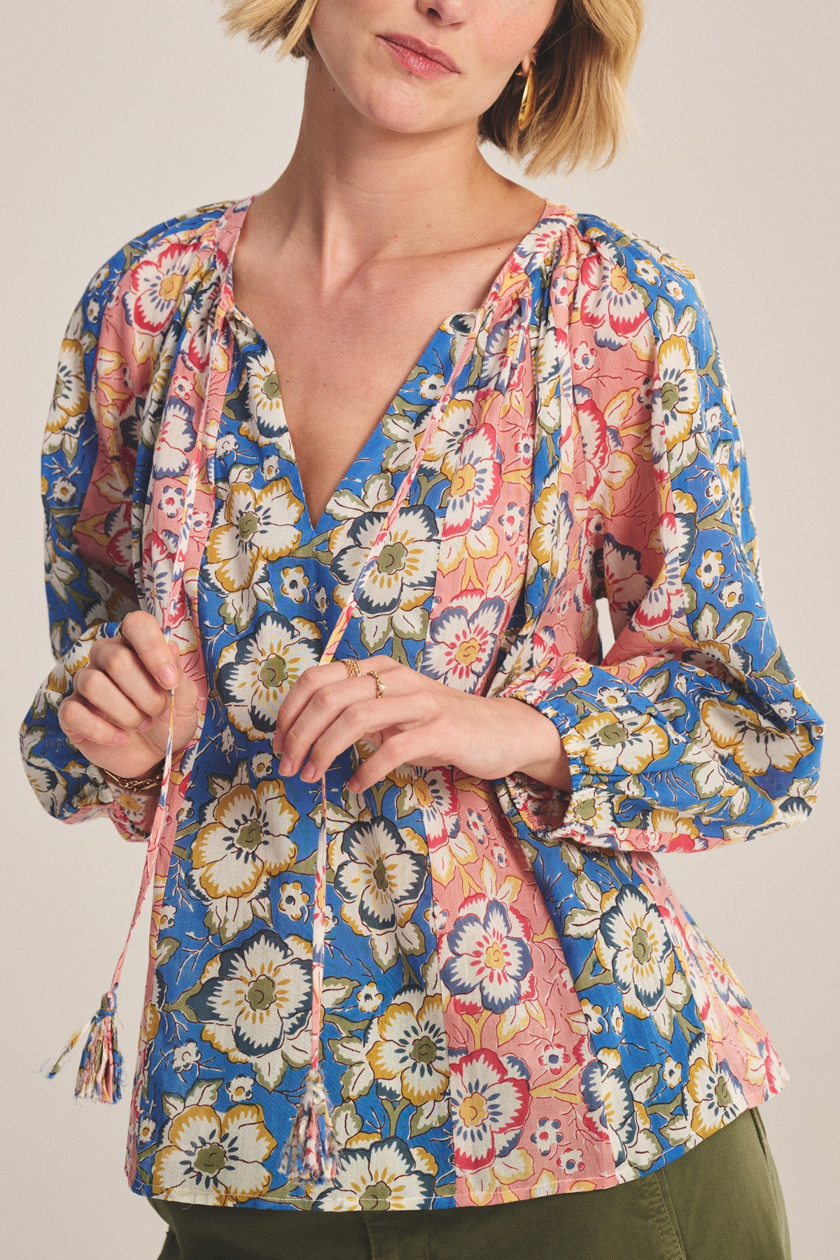 A woman wears the REESE TOP by Velvet by Graham & Spencer, a blue and pink floral V-neck blouse with puffed long sleeves, paired with olive green pants.