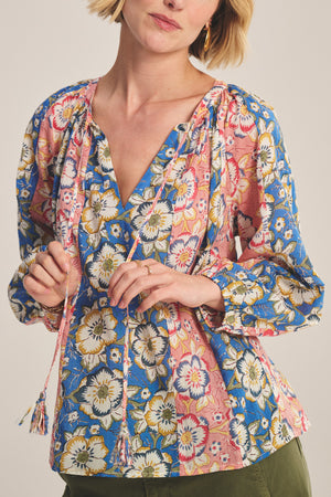 A woman wears the REESE TOP by Velvet by Graham & Spencer, a blue and pink floral V-neck blouse with puffed long sleeves, paired with olive green pants.