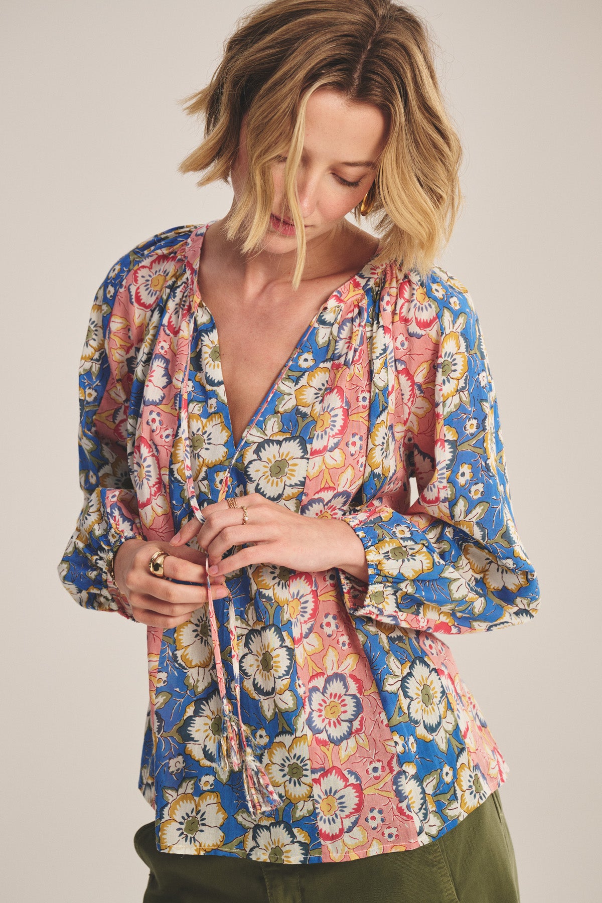 A woman with short blond hair wears the REESE TOP by Velvet by Graham & Spencer—a lightweight printed cotton blouse in blue, pink, and yellow with a V-neckline and long sleeves—as she adjusts the fabric ties at the neckline.-41008391848129