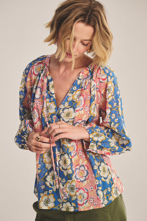 A woman with short blond hair wears the REESE TOP by Velvet by Graham & Spencer—a lightweight printed cotton blouse in blue, pink, and yellow with a V-neckline and long sleeves—as she adjusts the fabric ties at the neckline.