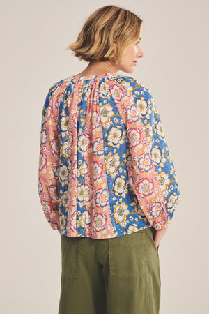 A person stands facing away, wearing the REESE TOP by Velvet by Graham & Spencer—a lightweight cotton blouse with pink and blue florals and puffed sleeves—paired with olive green pants.