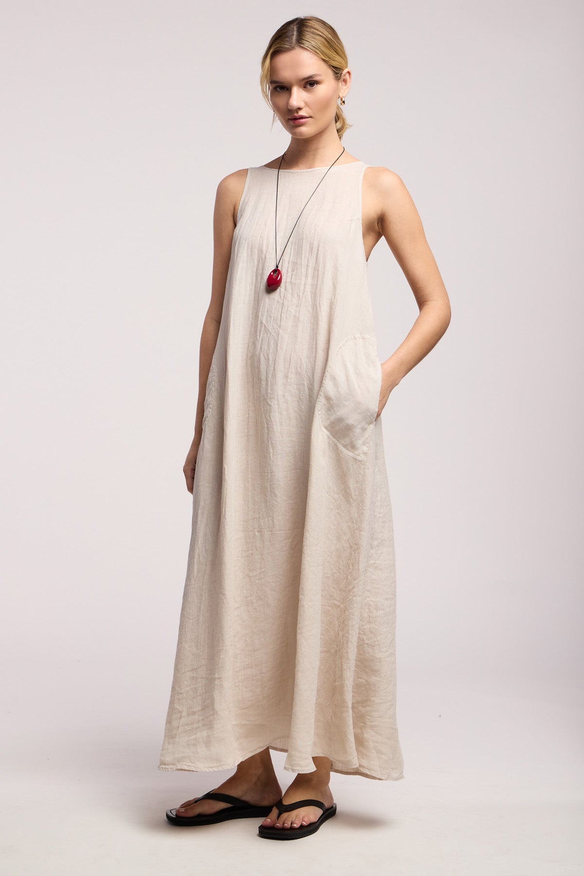 A woman in the RETA LINEN DRESS by Velvet by Graham & Spencer—sleeveless, beige, with side pockets—paired with black flip-flops and a long necklace featuring a red pendant stands against a plain light background; ideal for effortless summer style.