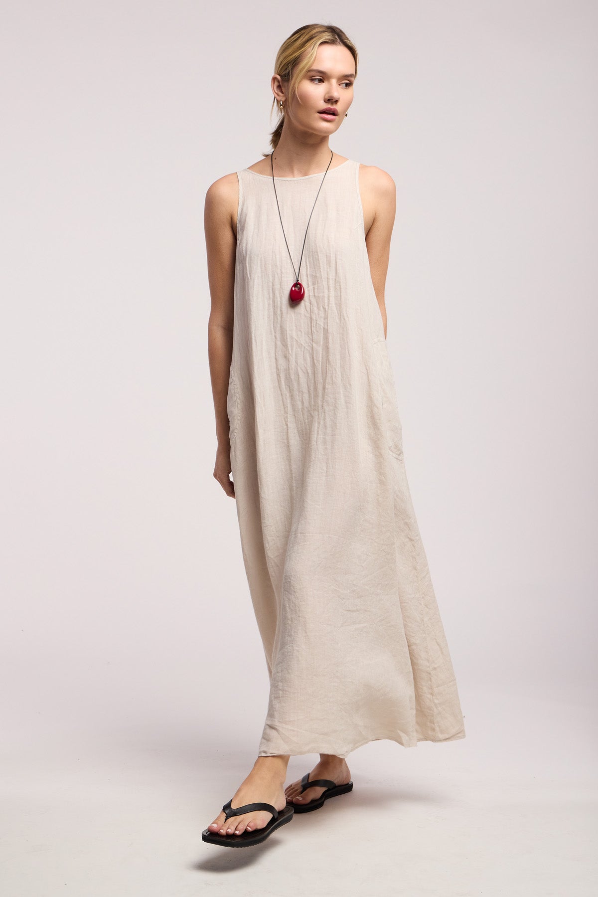 A woman wears the RETA LINEN DRESS by Velvet by Graham & Spencer—a sleeveless beige maxi styled with black sandals and a red pendant necklace, standing against a plain light background, ideal for warm-weather looks.