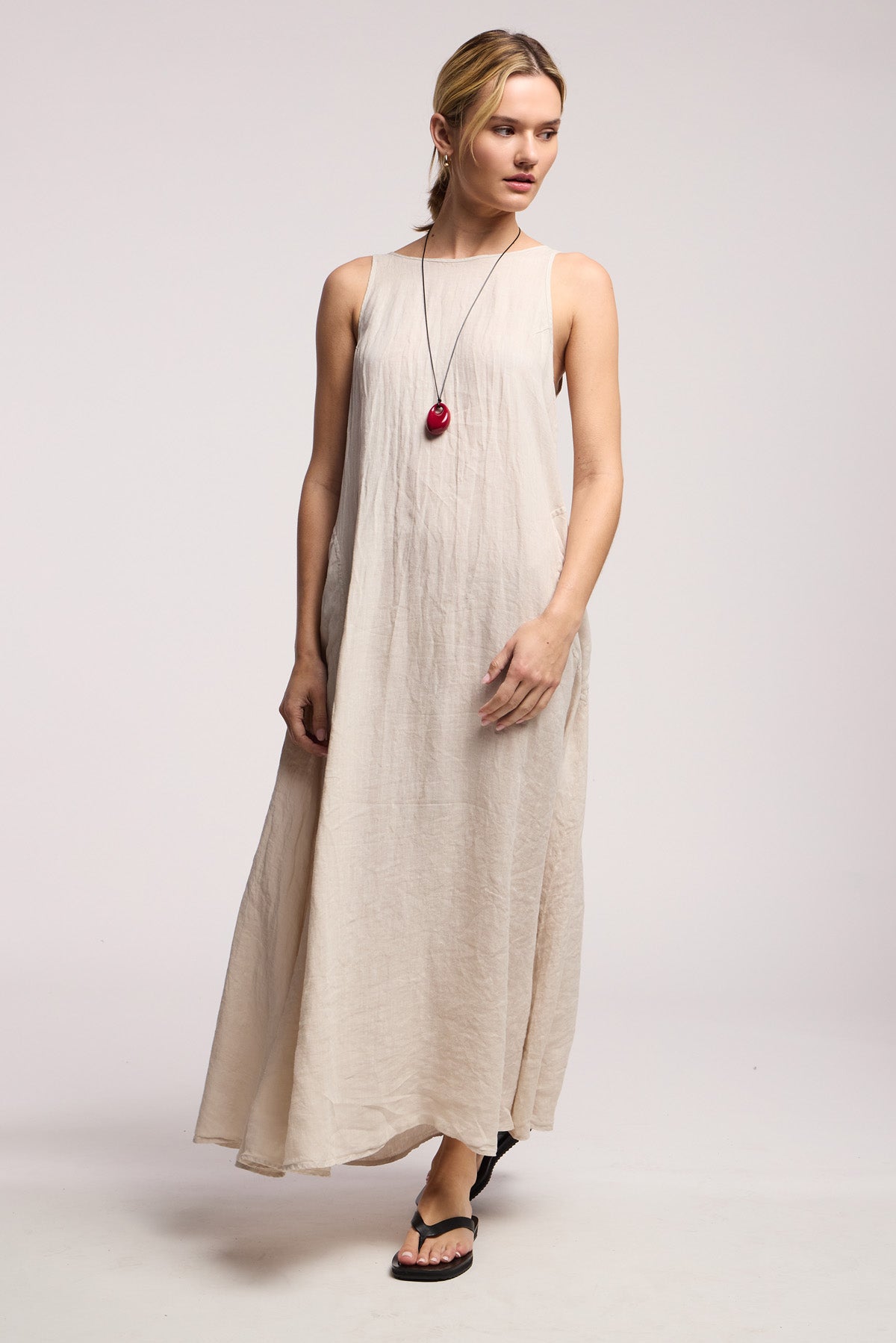 A person wearing the RETA LINEN DRESS by Velvet by Graham & Spencer in light beige, paired with a red pendant necklace and black sandals, stands against a plain light background.
