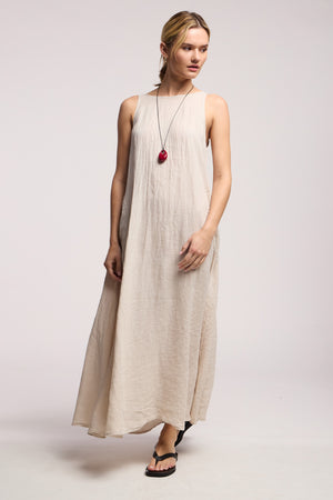 A person wearing the RETA LINEN DRESS by Velvet by Graham & Spencer in light beige, paired with a red pendant necklace and black sandals, stands against a plain light background.