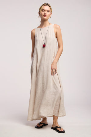 A person models the RETA LINEN DRESS by Velvet by Graham & Spencer, a sleeveless linen maxi with side pockets, paired with black flip-flops and a long red-pendant necklace against a plain background for a minimal look.