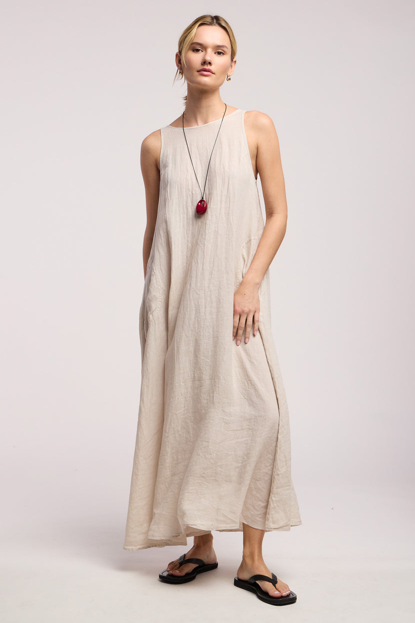 A person models the RETA LINEN DRESS by Velvet by Graham & Spencer, a sleeveless linen maxi with side pockets, paired with black flip-flops and a long red-pendant necklace against a plain background for a minimal look.