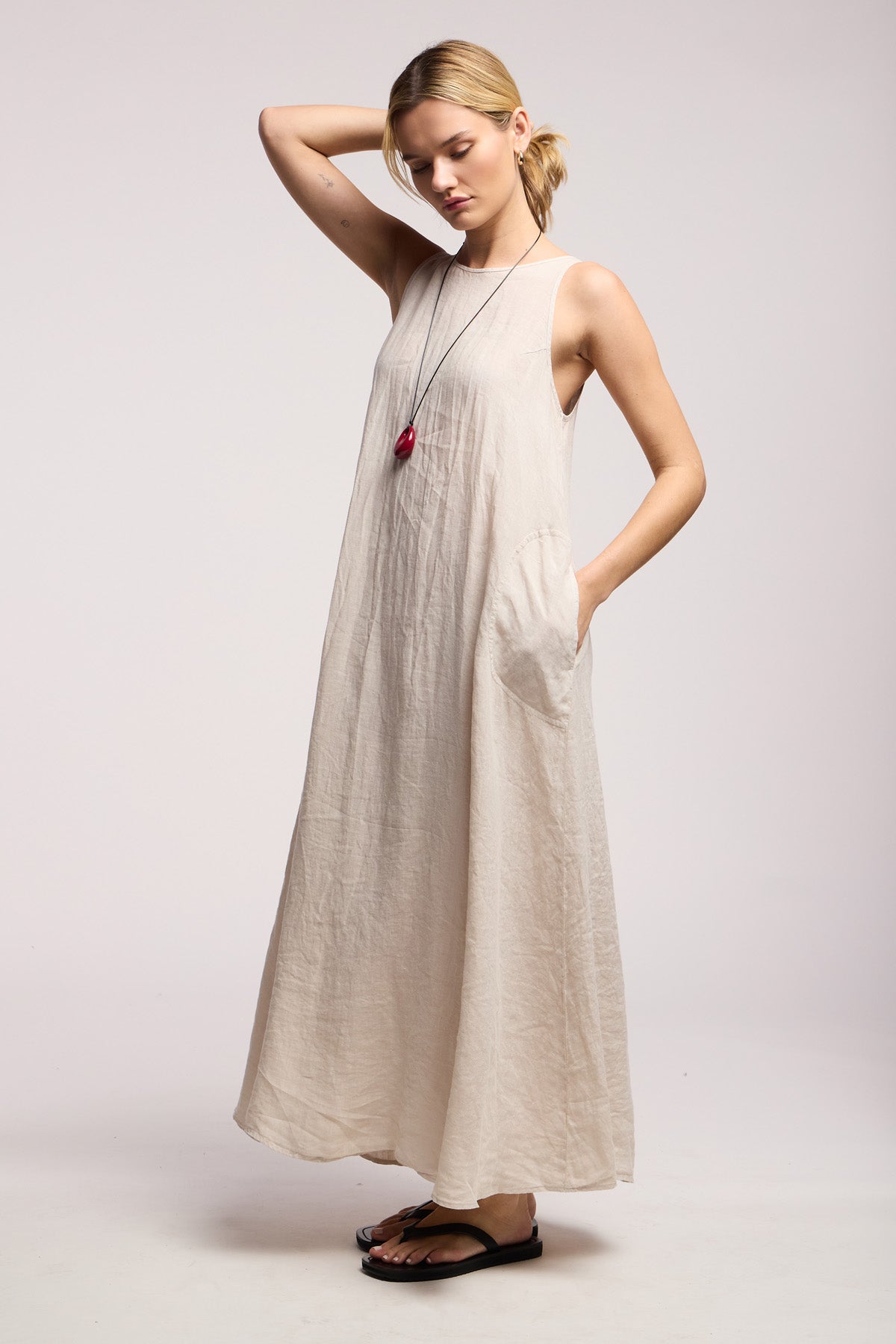 A woman wears the RETA LINEN DRESS by Velvet by Graham & Spencer—a sleeveless maxi dress with pockets—paired with black sandals and a red pendant necklace, standing against a plain background.-Hover Image