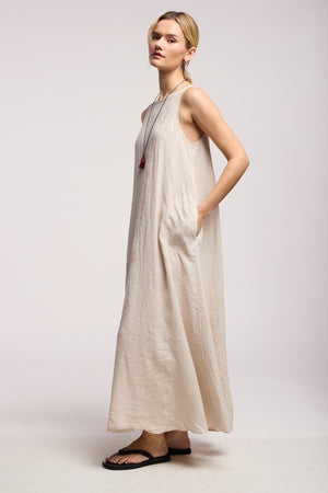 A woman stands in profile against a plain light background wearing the Velvet by Graham & Spencer RETA LINEN DRESS—a sleeveless maxi dress with side pockets—paired with black sandals and a long necklace for effortless warm-weather style.