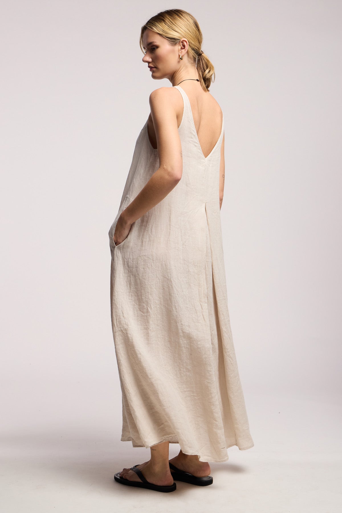 A woman stands sideways against a plain light background wearing the Velvet by Graham & Spencer RETA LINEN DRESS, a loose, sleeveless maxi with pockets, paired with black sandals—ideal for effortless warm-weather style.-41372106752193