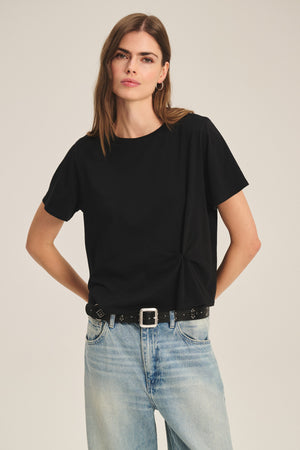 Wearing the RHOU TEE by Velvet by Graham & Spencer, tucked into light blue jeans with a black belt, this person stands hands on hips against a plain background.