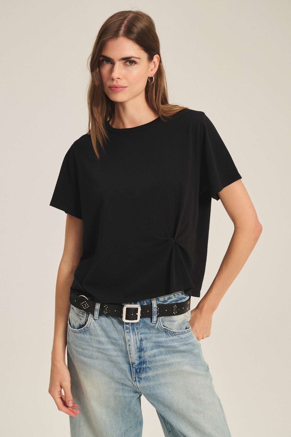 A woman wears the RHOU TEE by Velvet by Graham & Spencer—a black boxy soft cotton jersey—paired with light blue jeans and a black studded belt, standing against a plain background.