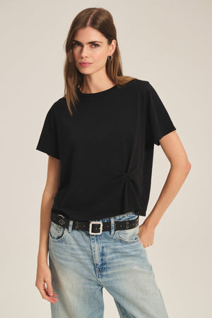 A woman wears the RHOU TEE by Velvet by Graham & Spencer—a black boxy soft cotton jersey—paired with light blue jeans and a black studded belt, standing against a plain background.