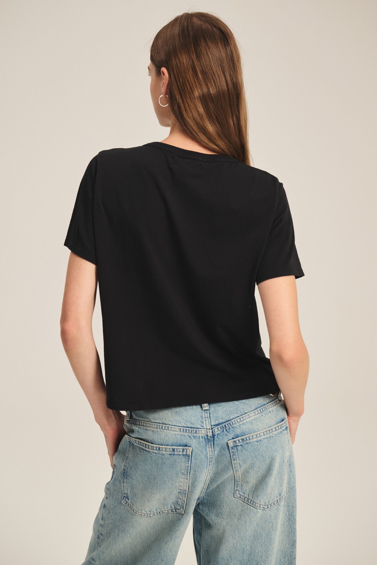 Someone with long brown hair is seen from behind wearing the Velvet by Graham & Spencer RHOU TEE in black cotton jersey and light blue jeans, standing with hands in their back pockets.-41467532476609