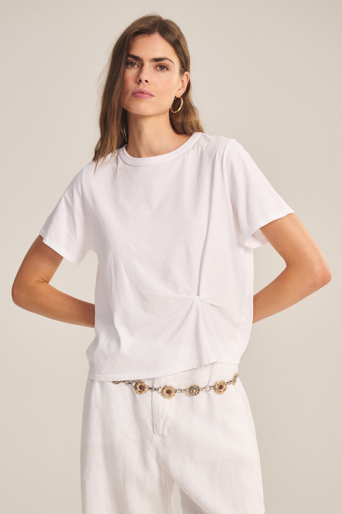 A woman wears the RHOU TEE by Velvet by Graham & Spencer—a relaxed fit, plain white cotton jersey boxy top with a subtle front knot—paired with white pants, hoop earrings, and a decorative chain belt.