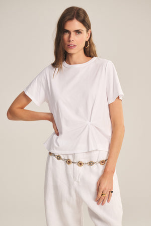 A woman wears the RHOU TEE by Velvet by Graham & Spencer—a white cotton jersey boxy top with a front twist—paired with white pants, a decorative belt, hoop earrings, and rings.