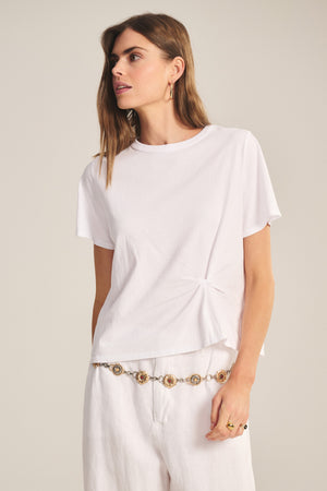 A woman models the Velvet by Graham & Spencer RHOU TEE, a relaxed fit white cotton jersey top with twist detail, paired with white pants and gold accessories, against a neutral background.