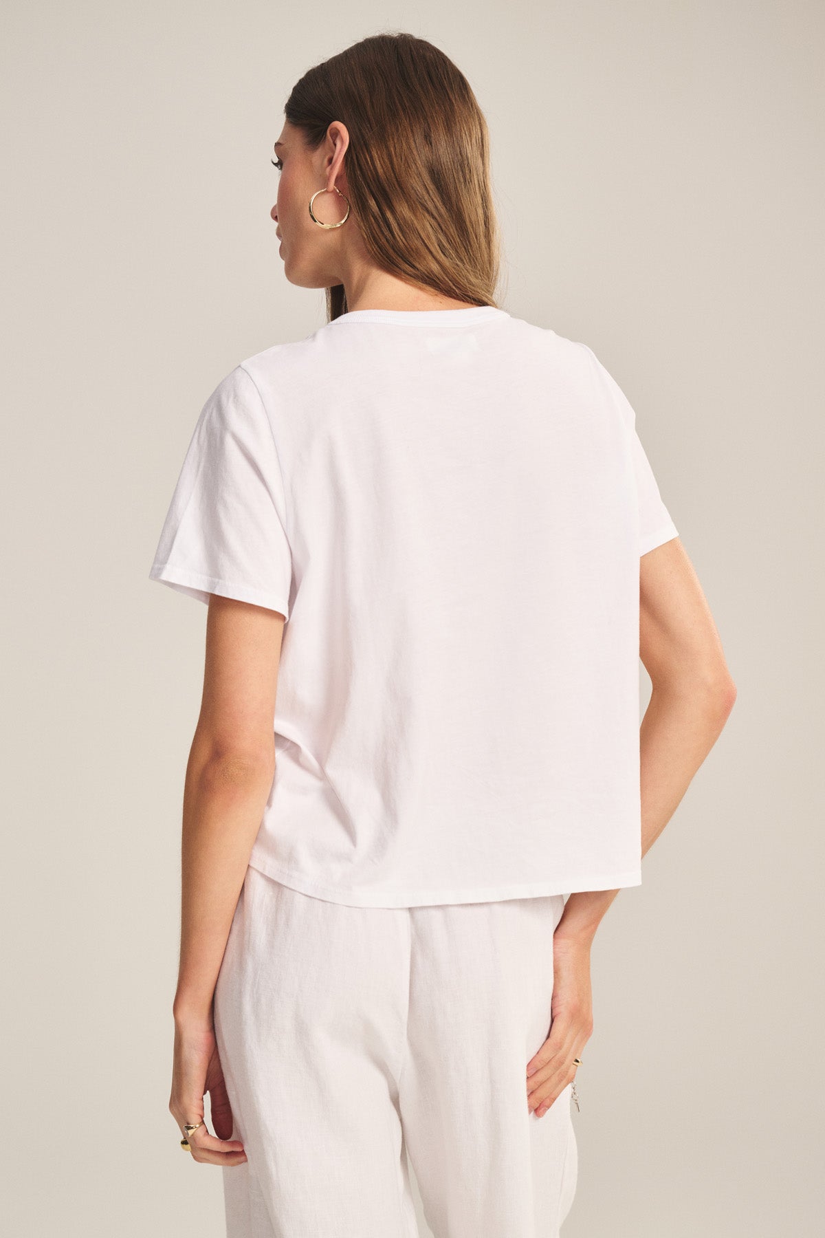 A woman with long brown hair models the RHOU TEE by Velvet by Graham & Spencer, a relaxed fit white cotton jersey short-sleeve t-shirt, paired with white pants and photographed from the back against a plain background.-41008394141889
