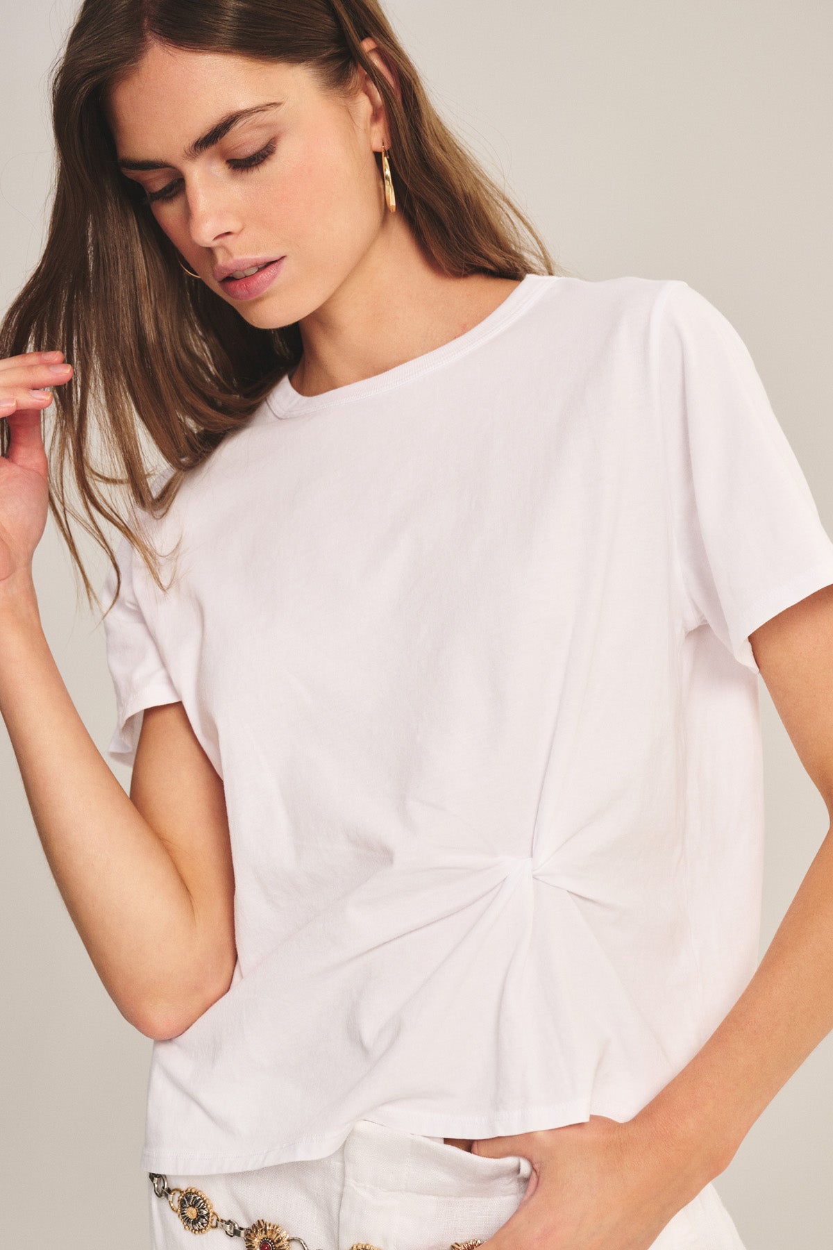 A woman in the Velvet by Graham & Spencer RHOU TEE, a relaxed fit white cotton jersey top with a front knot detail, pairs it with light-colored pants and gold hoops, looking downward with one hand raised.-41008394109121