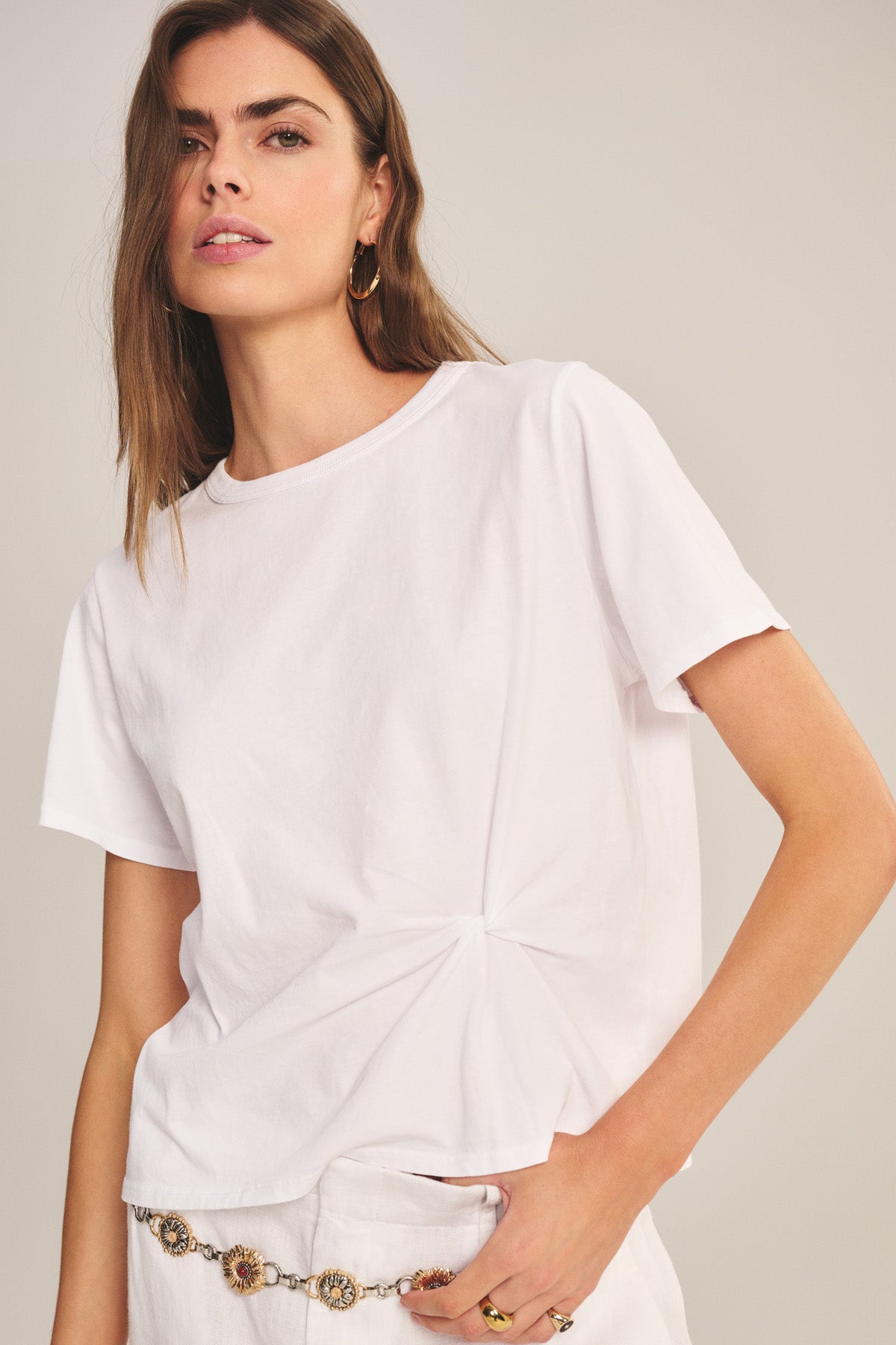 A woman wears the RHOU TEE by Velvet by Graham & Spencer—a white cotton jersey boxy tee with a twist detail at the waist—paired with white pants, standing against a plain background.