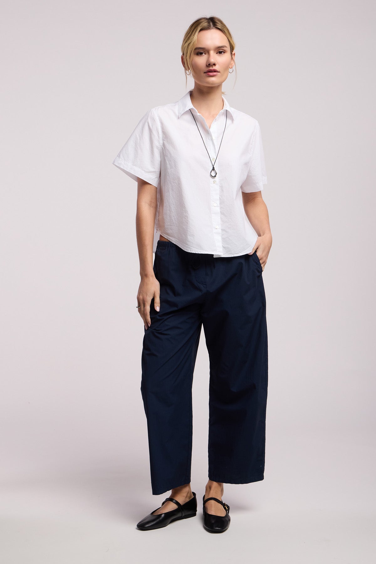 A person stands against a plain background, wearing the ALANA SHIRT by Velvet by Graham & Spencer—a white cotton poplin, short-sleeve shirt—paired with navy wide-leg pants, black shoes, a necklace, and hands in pockets.-41370843644097