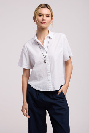 A woman models the ALANA SHIRT by Velvet by Graham & Spencer—short-sleeve cotton poplin—paired with dark pants and a necklace, standing with one hand in her pocket against a plain background.