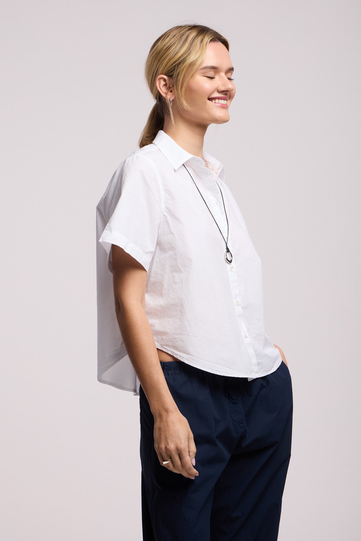 A woman stands sideways with hands in pockets, wearing the ALANA SHIRT by Velvet by Graham & Spencer—a white cotton poplin short-sleeve shirt—paired with navy pants and a pendant necklace, hair in a ponytail, against a neutral background.-41370843513025