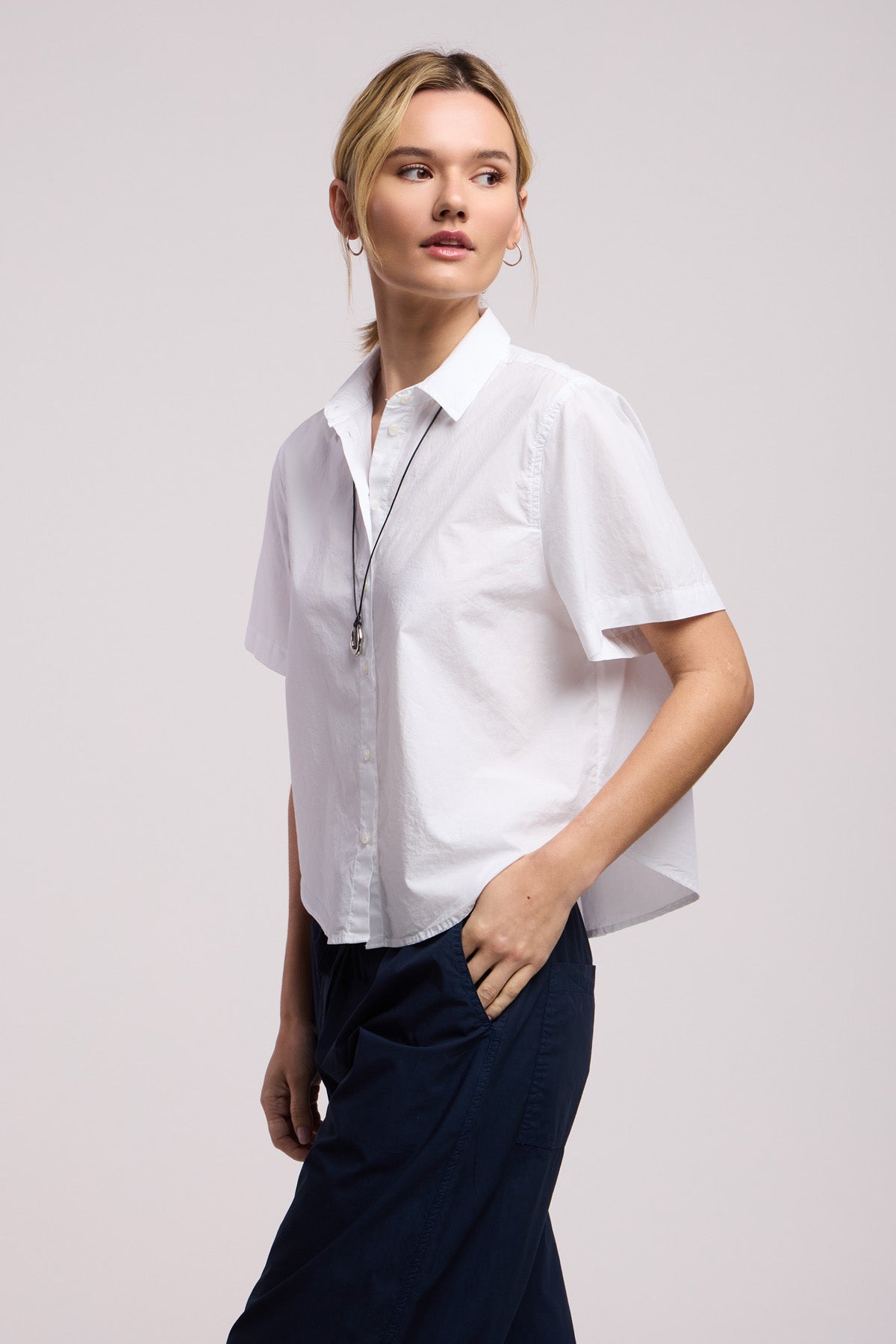 Model wears the ALANA SHIRT by Velvet by Graham & Spencer, a short-sleeve white cotton poplin top, paired with dark pants. She stands with one hand in her pocket, gazing slightly to the side against a plain background.