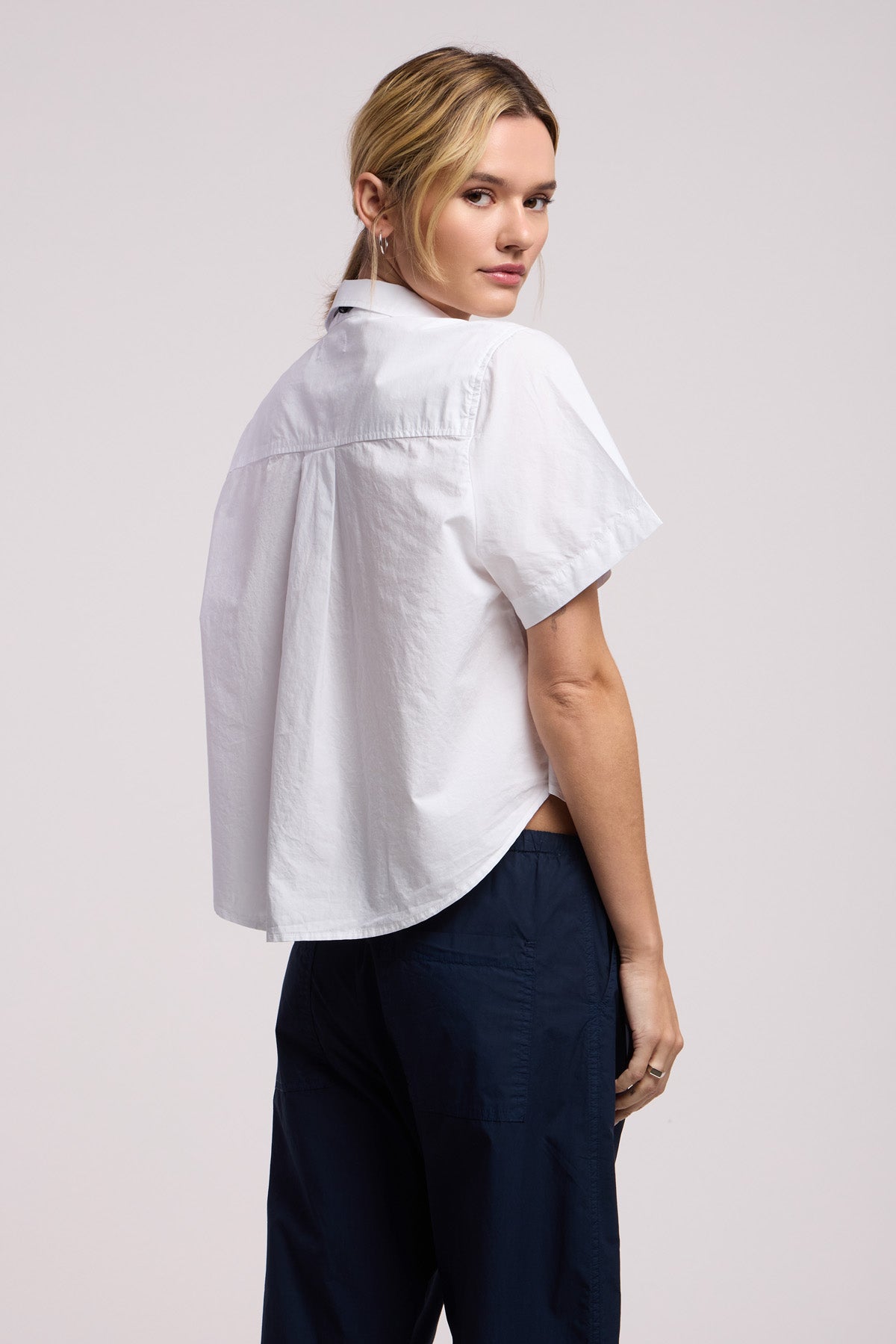 A woman faces away, looking over her shoulder, wearing the Velvet by Graham & Spencer ALANA SHIRT—a white cotton poplin short-sleeve top—paired with dark blue pants against a plain background.-41370843414721