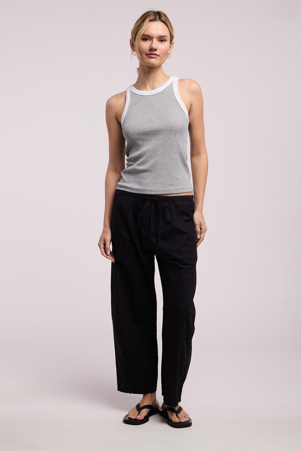 A person faces forward wearing the Velvet by Graham & Spencer ALLEGRA PANT—black cropped, barrel-leg pull-on pants—with a grey tank top with white trim and black flip-flops, set against a plain background.