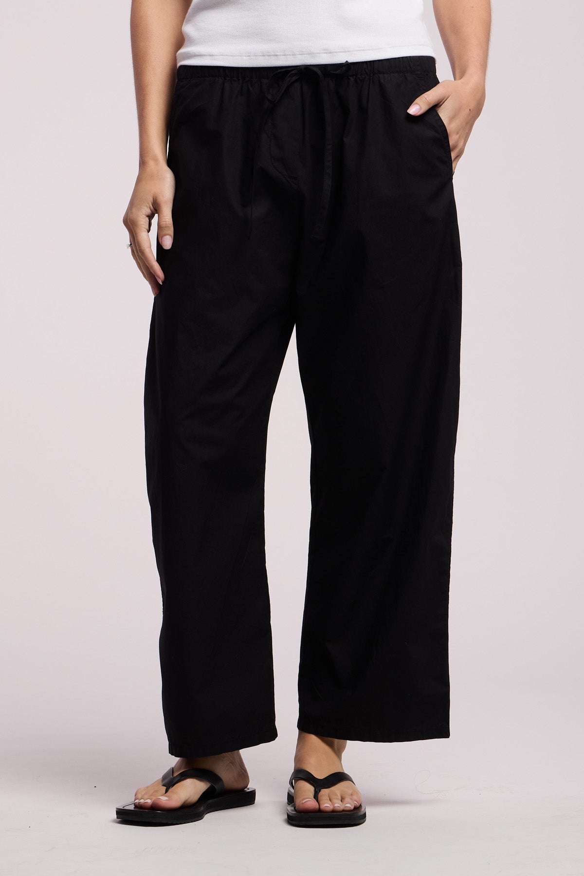 Person wearing the Velvet by Graham & Spencer ALLEGRA PANT in black, paired with a white top and black flip-flops, standing with one hand in their pocket against a plain background.-41370867138753