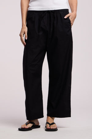 Person wearing the Velvet by Graham & Spencer ALLEGRA PANT in black, paired with a white top and black flip-flops, standing with one hand in their pocket against a plain background.