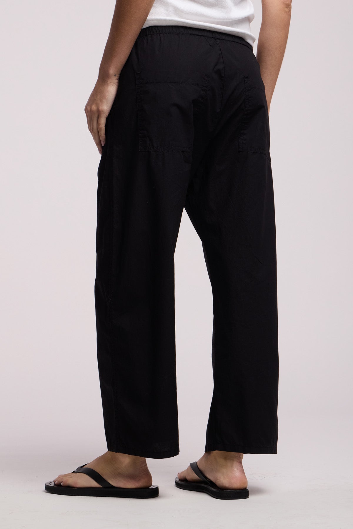 A person faces away from the camera, wearing Velvet by Graham & Spencer's ALLEGRA PANT—black lightweight cotton poplin pull-on pants with a cropped barrel leg—paired with a white top and black flip-flops against a plain background.