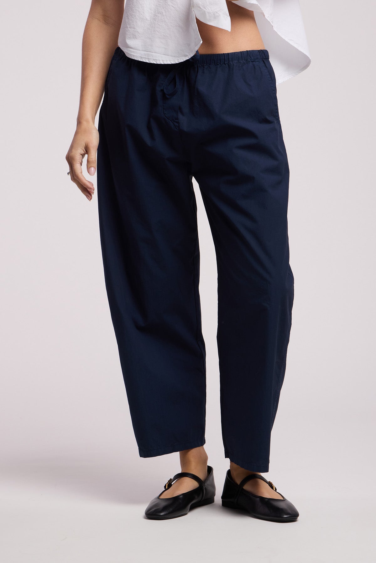 A person wears the ALLEGRA PANT by Velvet by Graham & Spencer—navy barrel-leg, cropped, lightweight cotton poplin—paired with a white top and black flats, standing against a light background.