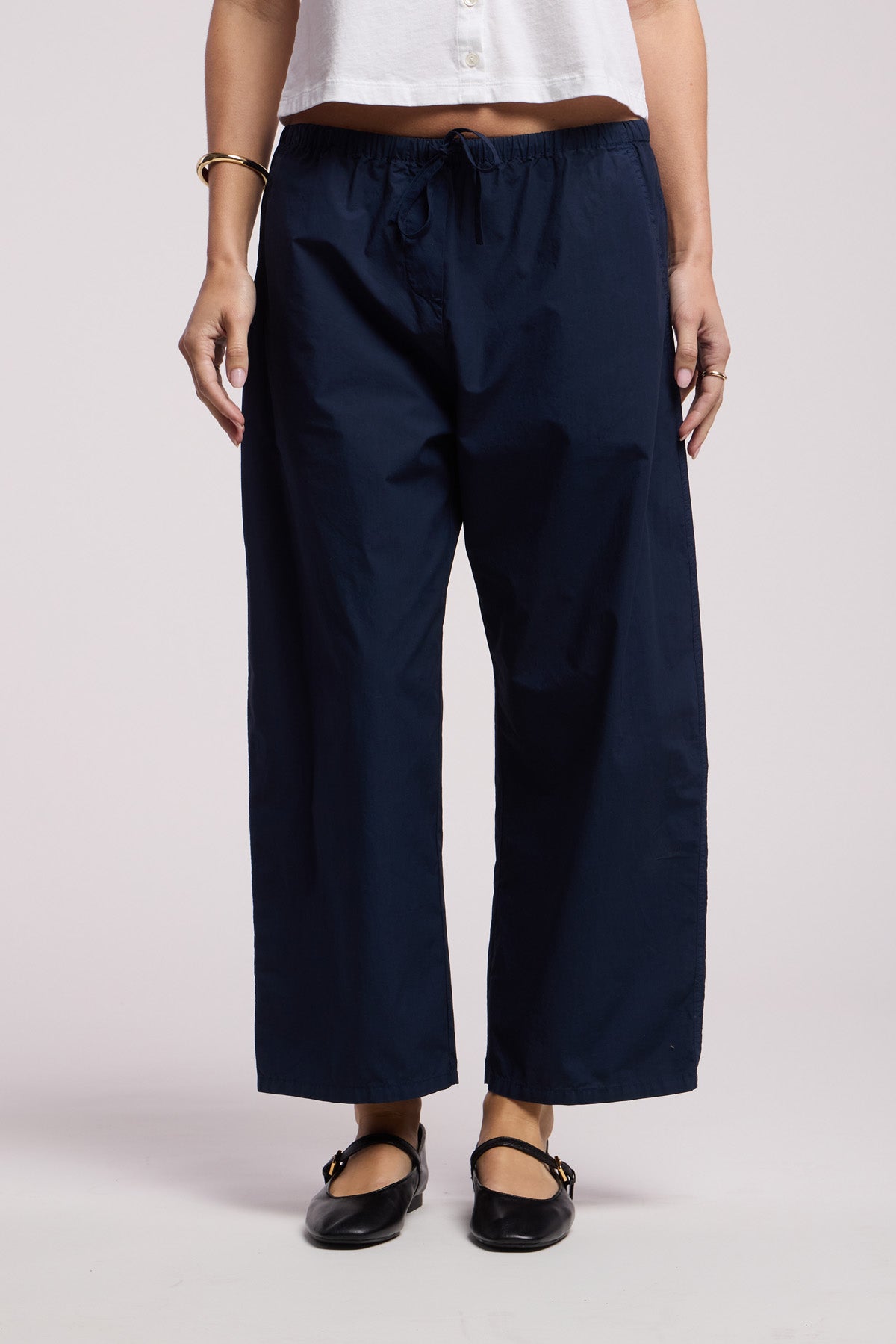 Wearing Velvet by Graham & Spencer's ALLEGRA PANT in navy blue cotton poplin with a white cropped top, black flat shoes, bracelets, and rings, someone stands against a plain background.-41527309009089