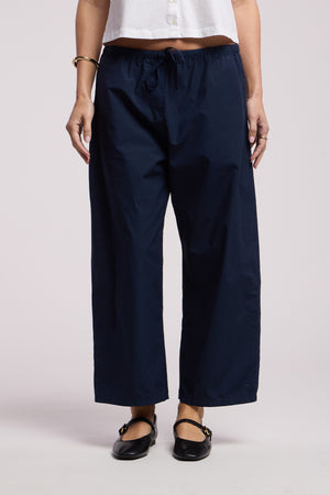 Wearing Velvet by Graham & Spencer's ALLEGRA PANT in navy blue cotton poplin with a white cropped top, black flat shoes, bracelets, and rings, someone stands against a plain background.