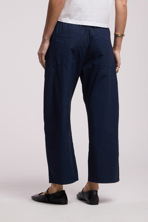 Person wearing a white lightweight cotton poplin top, ALLEGRA PANT by Velvet by Graham & Spencer in navy blue with a cropped barrel-leg fit, black flats, and a gold bracelet, standing with their back to the camera.
