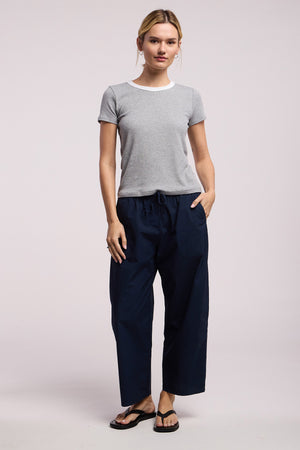 A person faces forward, wearing the Velvet by Graham & Spencer MILEY TEE—a grey short-sleeve fitted crewneck—paired with loose dark blue pants and black flip-flops against a plain background.