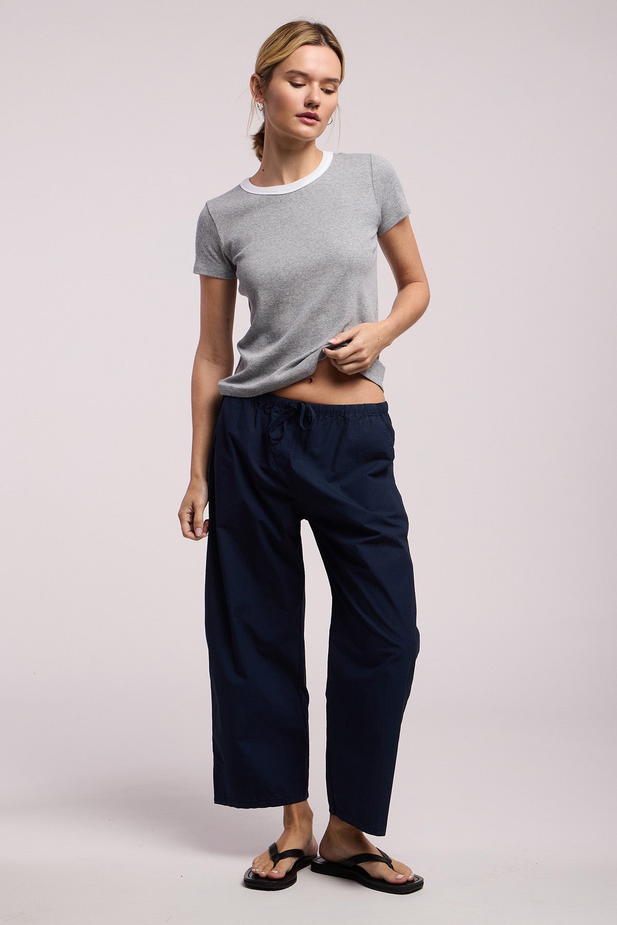 A person stands against a plain background wearing the ALLEGRA PANT by Velvet by Graham & Spencer, featuring cropped barrel legs and a drawstring waist, paired with a grey short-sleeve t-shirt and black flip-flops.
