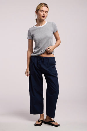 A person stands against a plain background wearing the ALLEGRA PANT by Velvet by Graham & Spencer, featuring cropped barrel legs and a drawstring waist, paired with a grey short-sleeve t-shirt and black flip-flops.