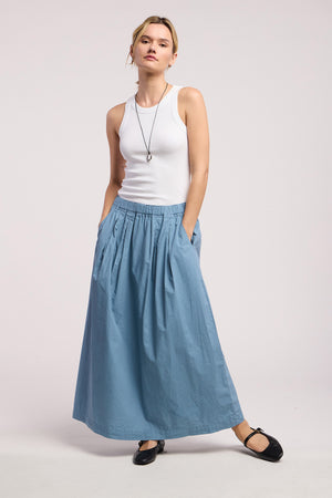 A woman stands against a plain background wearing Velvet by Graham & Spencer's CHESSA SKIRT—a blue cotton poplin maxi with pockets and an elastic waist—paired with a white sleeveless top, black ballet flats, and a pendant necklace.